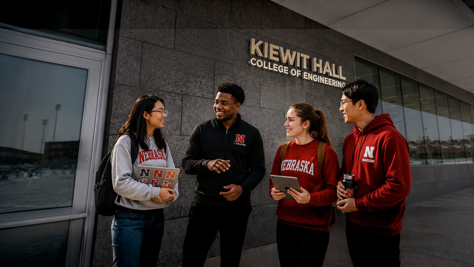Students smiling and talking outside Kiewit Hall, College of Engineering, wearing Nebraska attire. Image is AI generated.